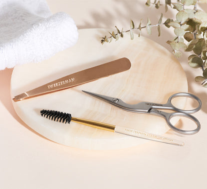 Tweezerman Eyebrow grooming tools including scissors, tweezers, and a brush on a rose quartz plate with a white towel and eucalyptus leaves.