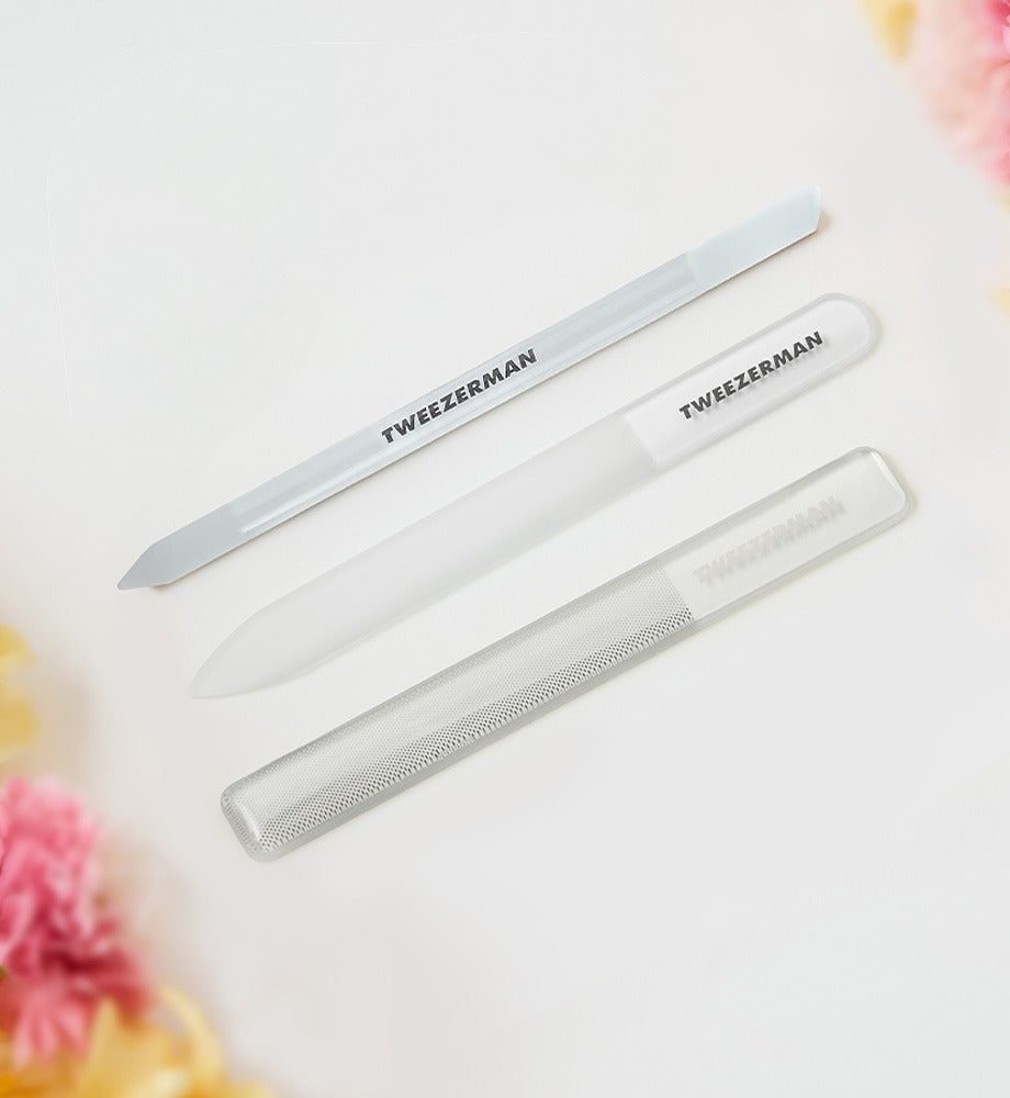 Tweezerman glass nail files and buffers on a white background with blurred flowers in the corner.