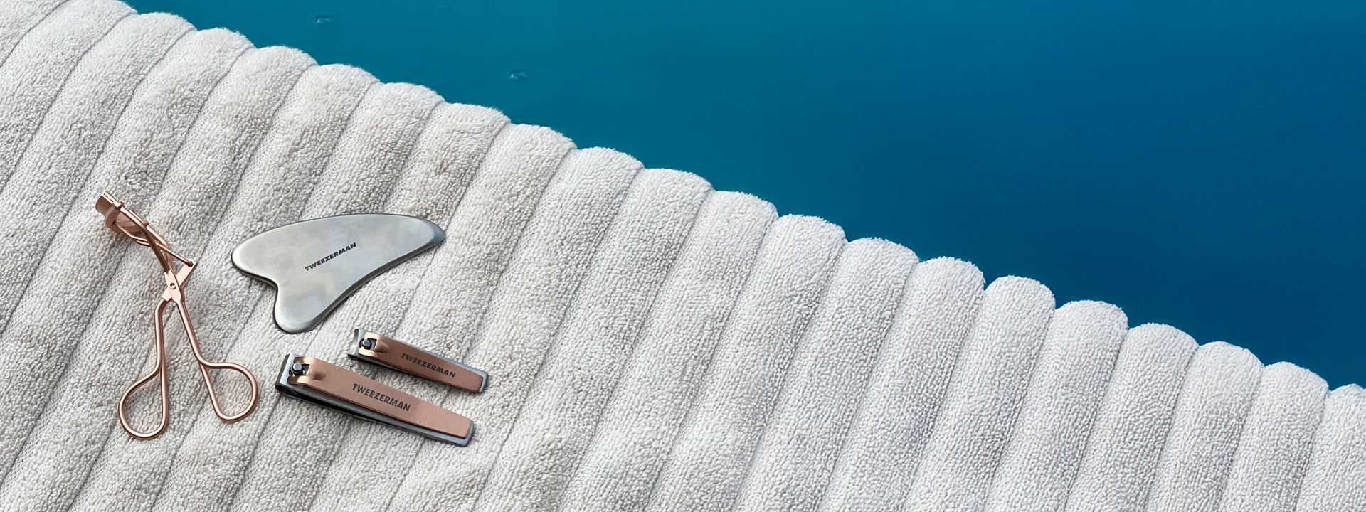 The Gua Sha, Eyelash Curler and Rose Gold Combo Clipper Set on a towel near a pool
