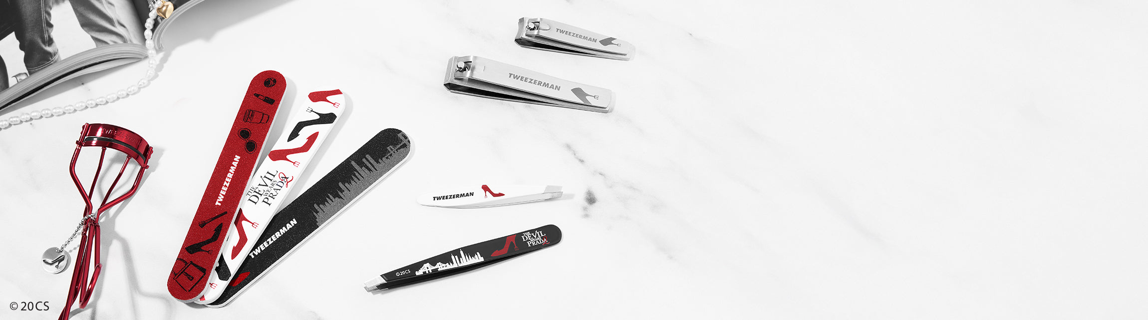 Set of beauty tools including a red eyelash curler, nail clippers and other The Devil Wears Prada branded tools on a white background