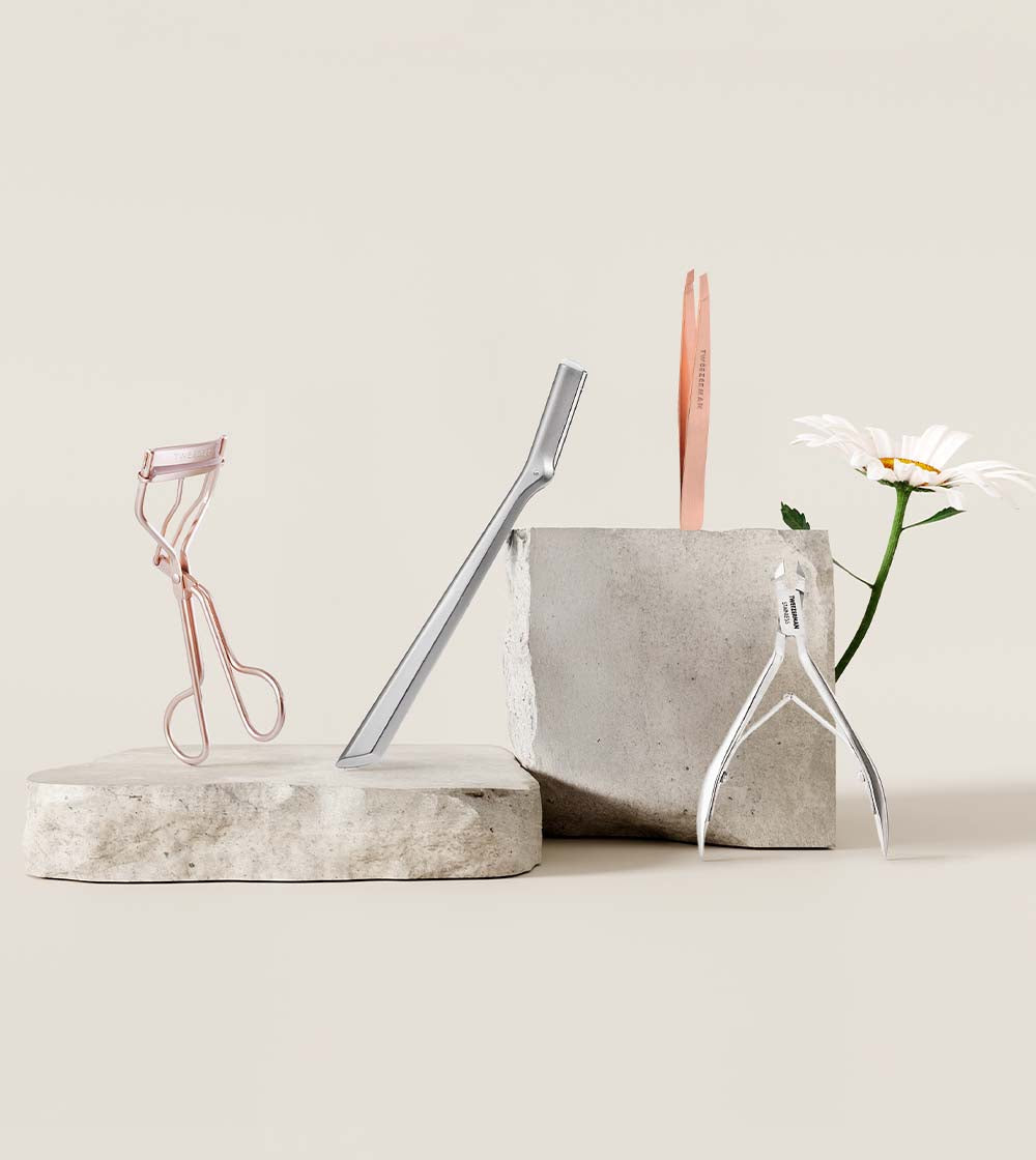 Beauty tools including a tweezer, eyelash curler, facial razor and cuticle nipper leaning against 2 concrete stands on a light surface.