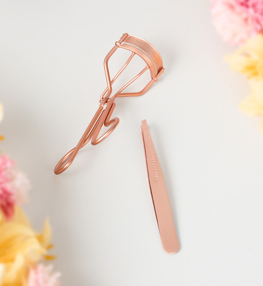 Rose gold eyelash curler and tweezers on a light background with floral elements.