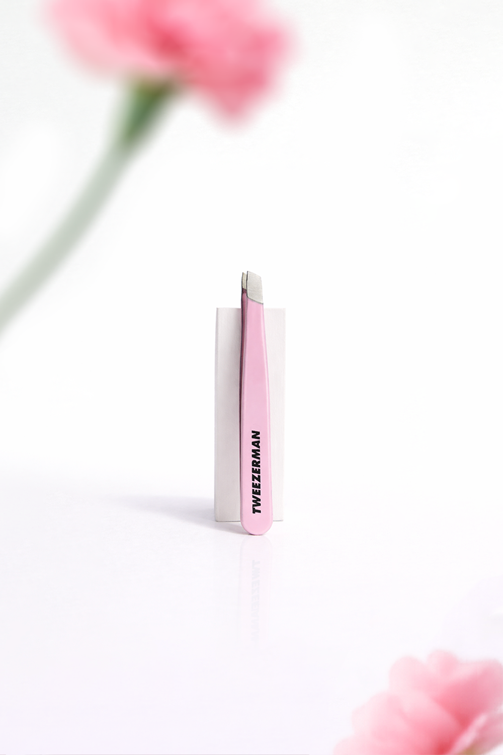 Pink micro mini tweezer with 'Tweezerman' branding on a white background with blurred pink flowers.