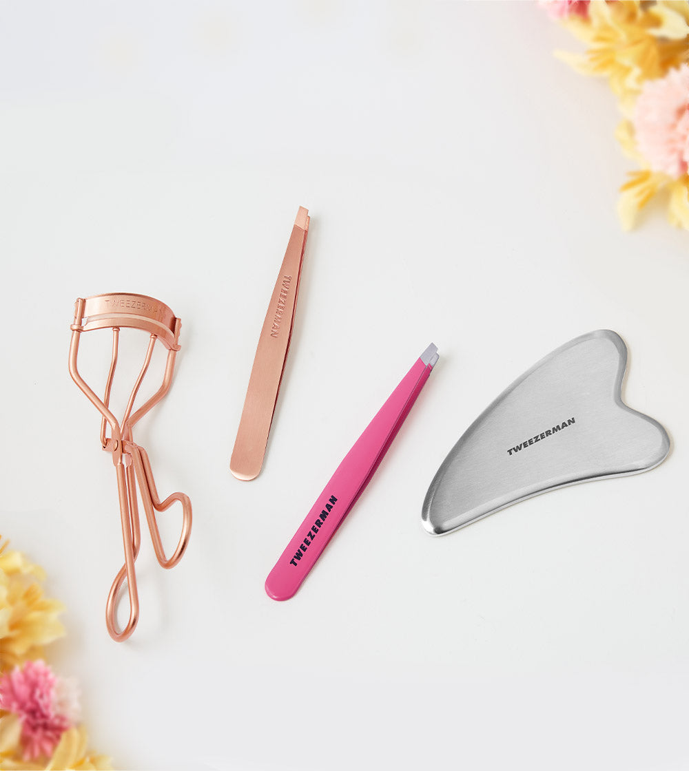 Set of beauty tools including a rose gold eyelash curler, pink tweezer, and silver gua sha tool on a white background with flowers.