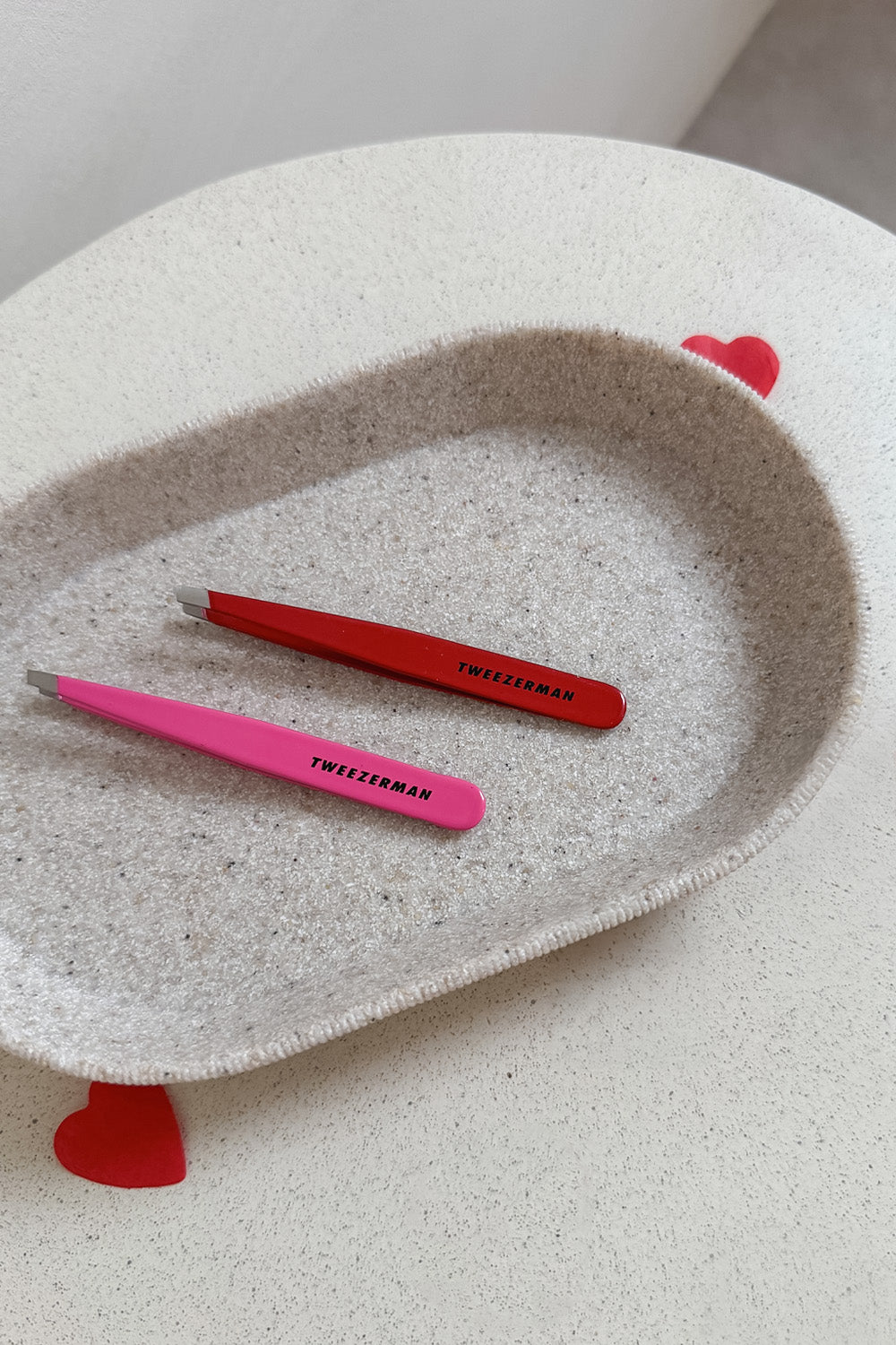 The Signature Red and the Neon Pink Slant Tweezers on a bathroom tray, with 2 hearts around it